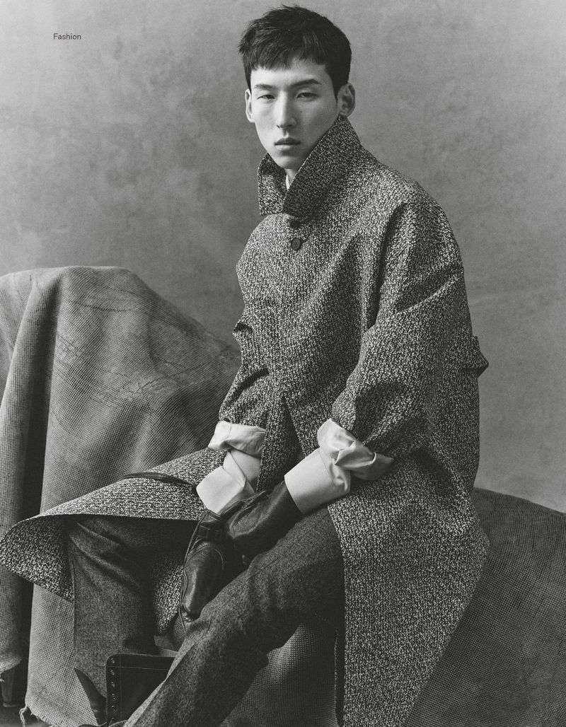 Portrait of a stylish Asian man in a wool coat and tailored pants, posing indoors against a mottled grey backdrop in soft studio light.