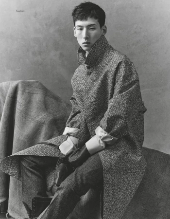 Portrait of a stylish Asian man in a wool coat and tailored pants, posing indoors against a mottled grey backdrop in soft studio light.