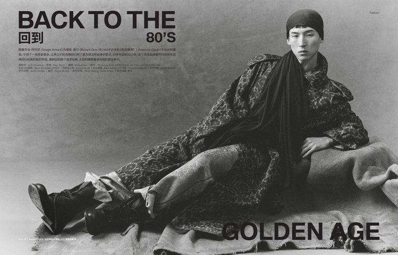 Model wearing a patterned coat and dark cap reclining on draped fabric against a textured grey background.