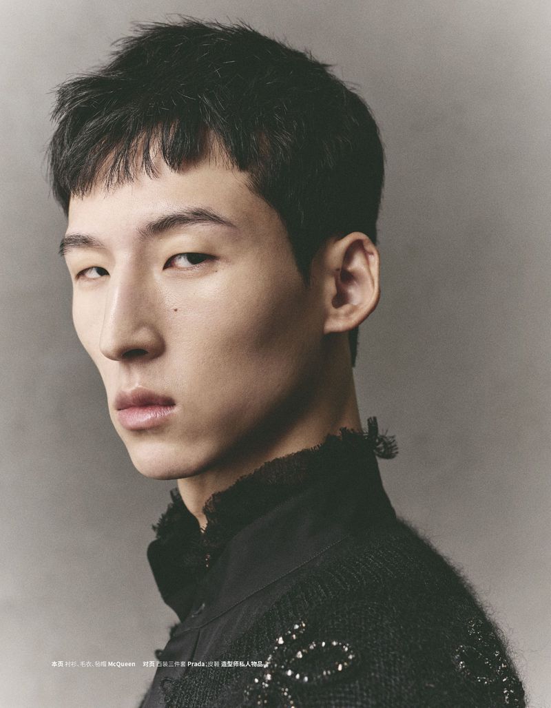 Close-up portrait of a young adult man in black high-necked shirt and embellishments against a muted grey studio backdrop. Fashion editorial.