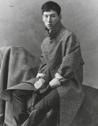 Monochrome fashion editorial portrait of a seated male model in a patterned trench coat against a textured studio backdrop