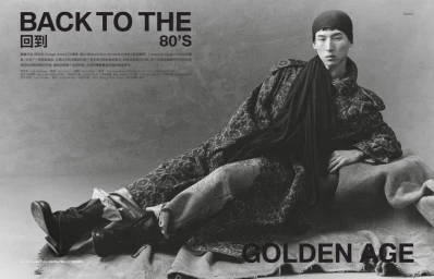 Model wearing a patterned coat and dark cap reclining on draped fabric against a textured grey background.