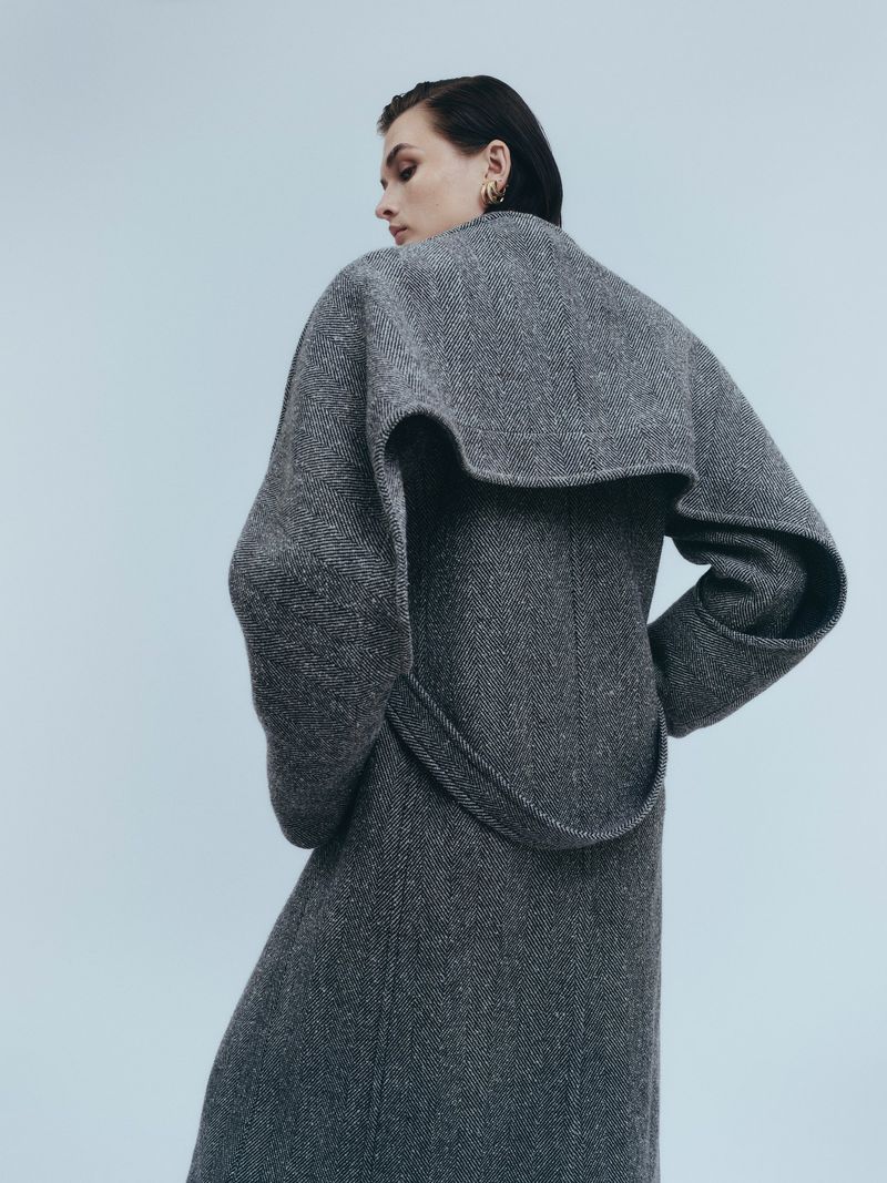 Woman poses in a gray wool coat against a pale blue background. Fashionable outerwear and neutral backdrop create a stylish look.