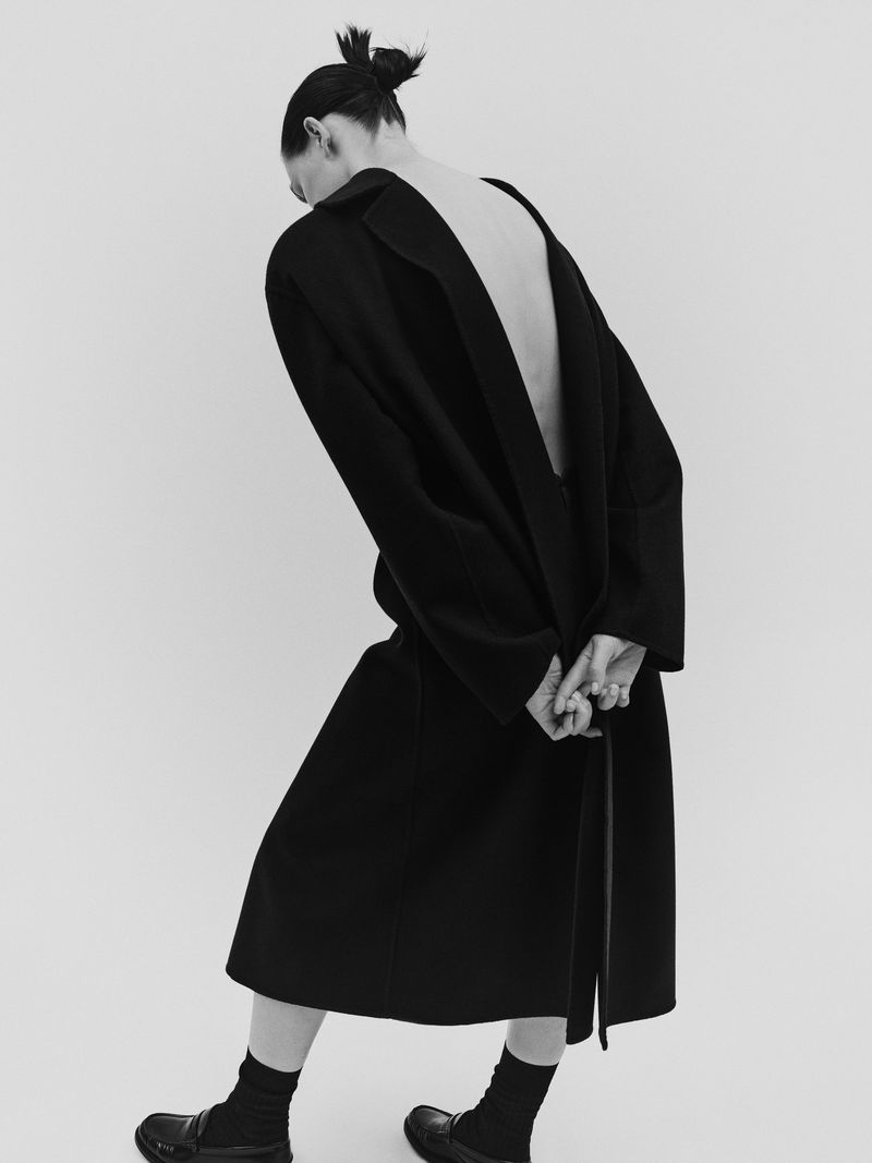 Woman in long black coat and dark socks stands against a light backdrop, revealing her back in this fashion shot. 