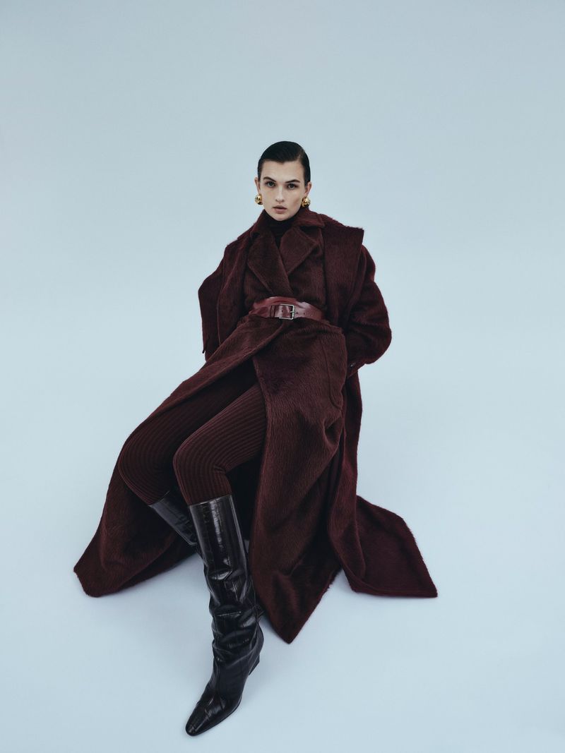 Fashion shot of a young adult woman in a long burgundy coat, leggings, and black boots, posed against a light blue backdrop.