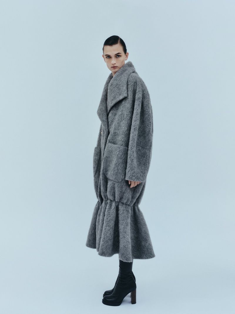 Fashion portrait of a female model wearing a voluminous grey coat with a ruffled hem and black boots in a light studio setting.