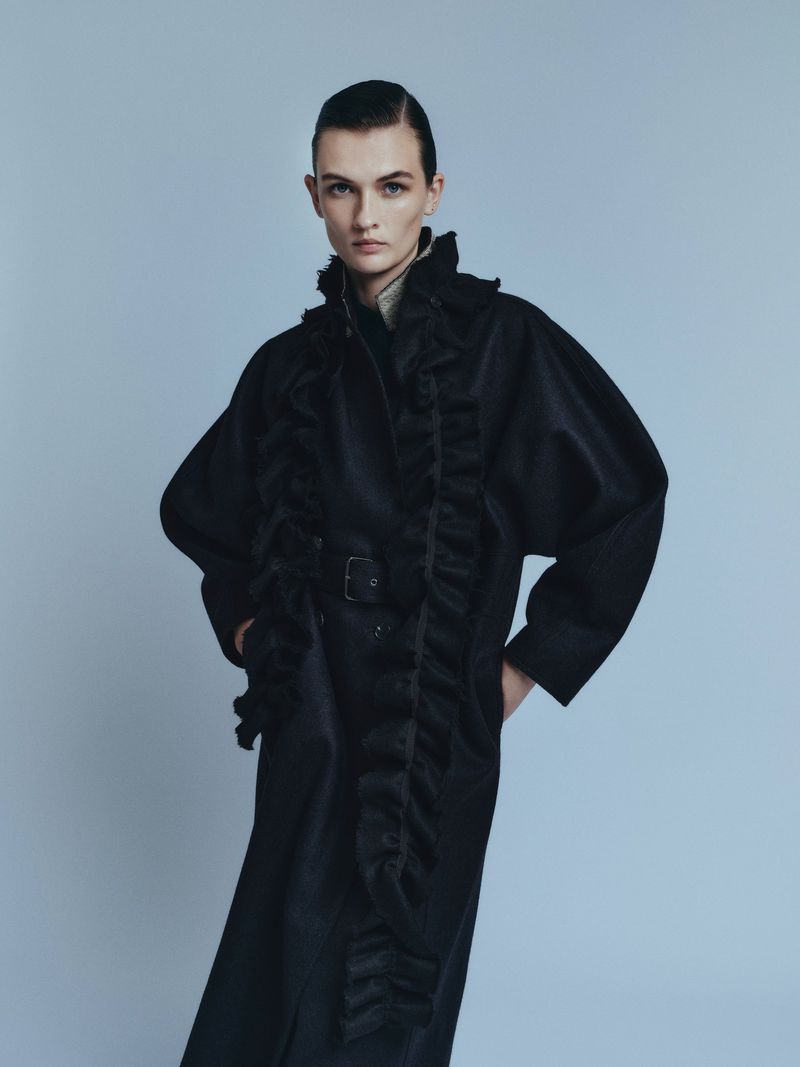 Fashion model poses in a long black belted coat with ruffles against a pale blue backdrop in a studio fashion shot.