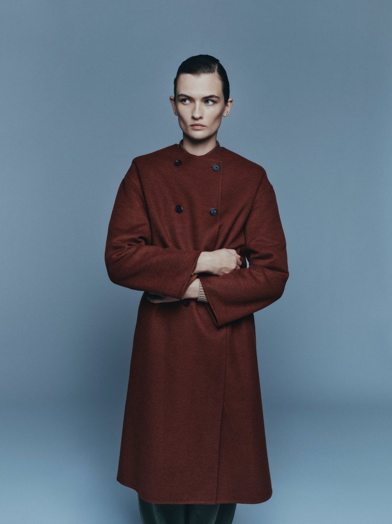 Studio portrait of a person with dark hair, wearing a long brown coat, standing with arms crossed against a light blue backdrop.