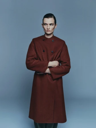 Studio portrait of a person with dark hair, wearing a long brown coat, standing with arms crossed against a light blue backdrop.