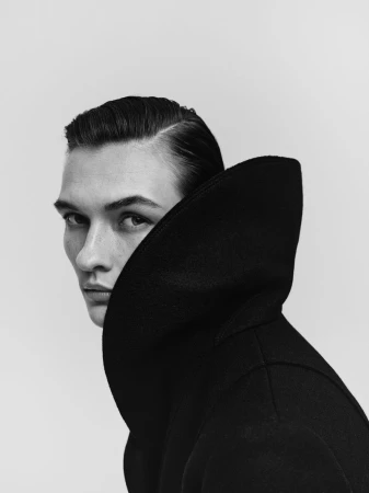 Monochrome portrait of a poised adult male wearing a dark wool coat with a large collar against a bright neutral backdrop.