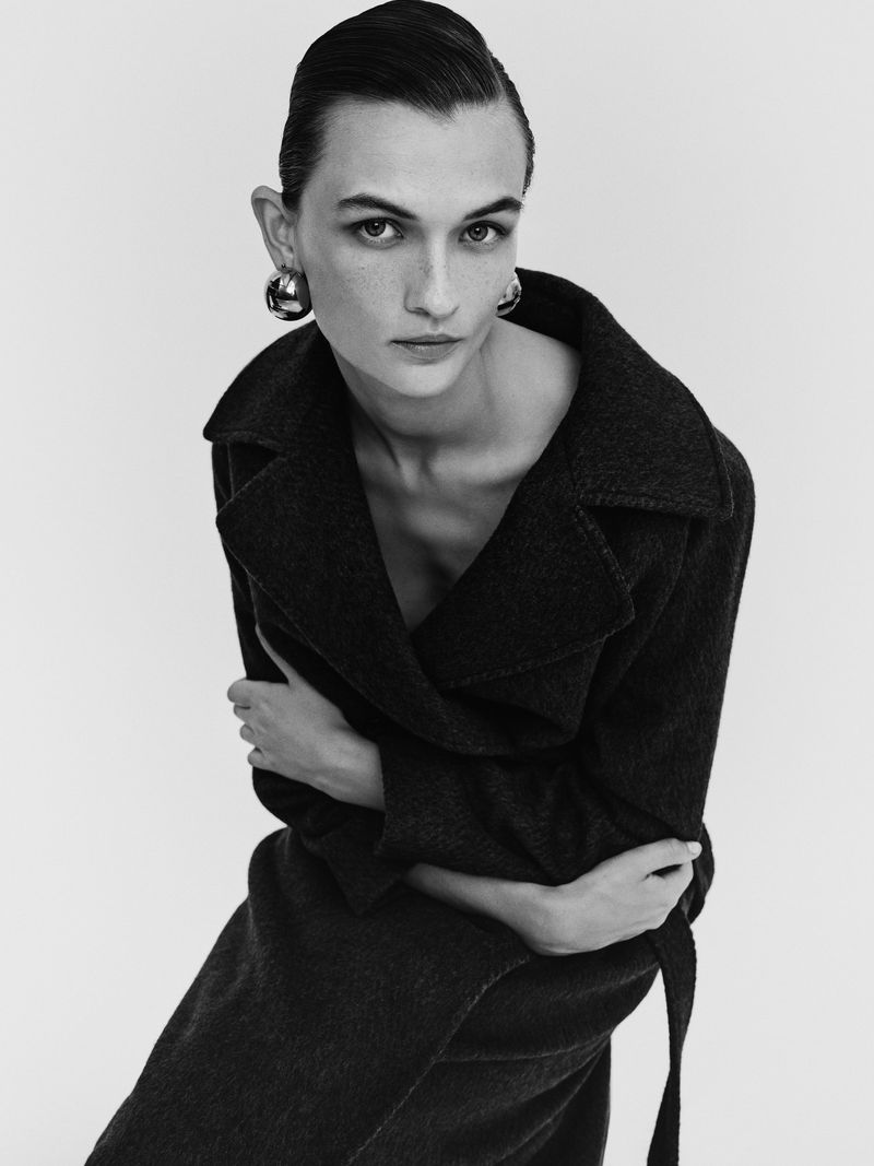 Monochrome portrait of a woman with arms crossed wearing a dark, belted coat and hoop earrings against a white background.