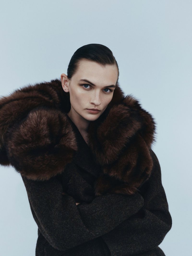 Woman wearing a dark grey coat with brown fur collar in a studio portrait with a blue background, arms crossed, looking at the viewer.