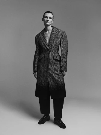 Model Lara Mullen posing in a tailored grey herringbone coat with an ombre hem, black trousers, and shoes against a grey studio backdrop.