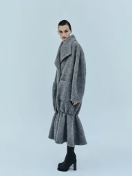 Model Lara Mullen posing in an oversized textured grey coat with flared hem and black heeled boots against a pale blue backdrop.