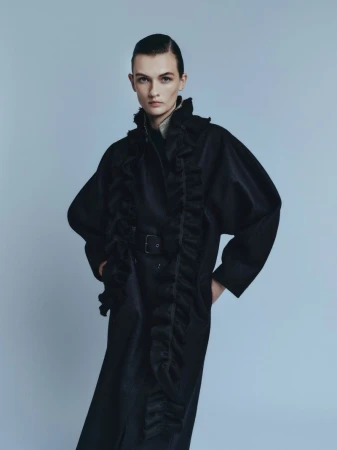 Model wearing a black ruffled coat against a plain neutral backdrop for the Amica project Il Cappotto