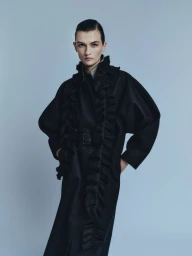 Model wearing a black ruffled coat against a plain neutral backdrop for the Amica project Il Cappotto