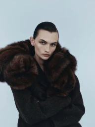 Model Lara Mullen wearing a dark coat with a voluminous brown fur collar against a light studio backdrop.