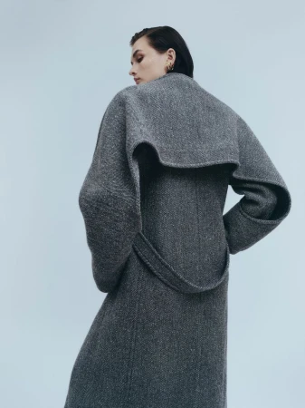 Model Lara Mullen wearing a grey tweed coat with a dramatic cape collar against a plain studio background.