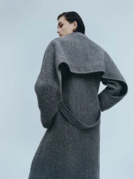 Model Lara Mullen wearing a grey tweed coat with a dramatic cape collar against a plain studio background.