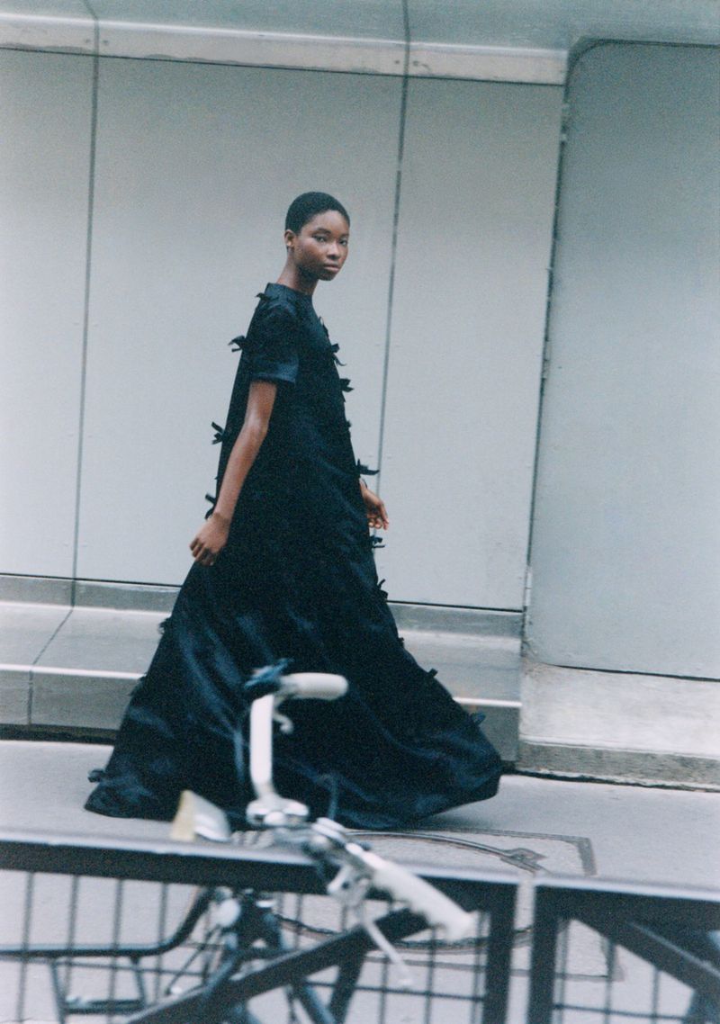 A lone model in a black dress adorned with bows walks along an outdoor city sidewalk, a bicycle parked nearby adding to the urban scene.
