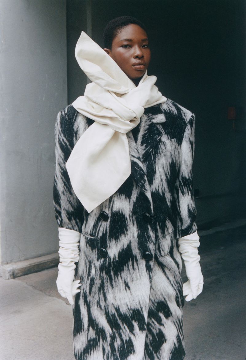 A Black female fashion model poses in an urban setting wearing a black and white coat with a white scarf and gloves.