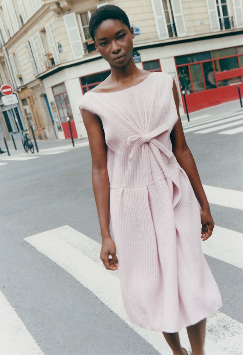 Black female model wearing a pale pink sleeveless pleated dress with a front knot detail standing in an urban pedestrian crosswalk.
