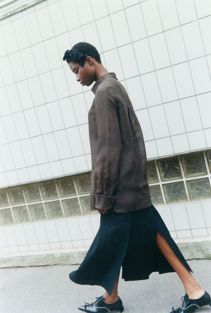A fashion shot of an African American woman in a brown top and black skirt walking in front of a white tiled wall and concrete.