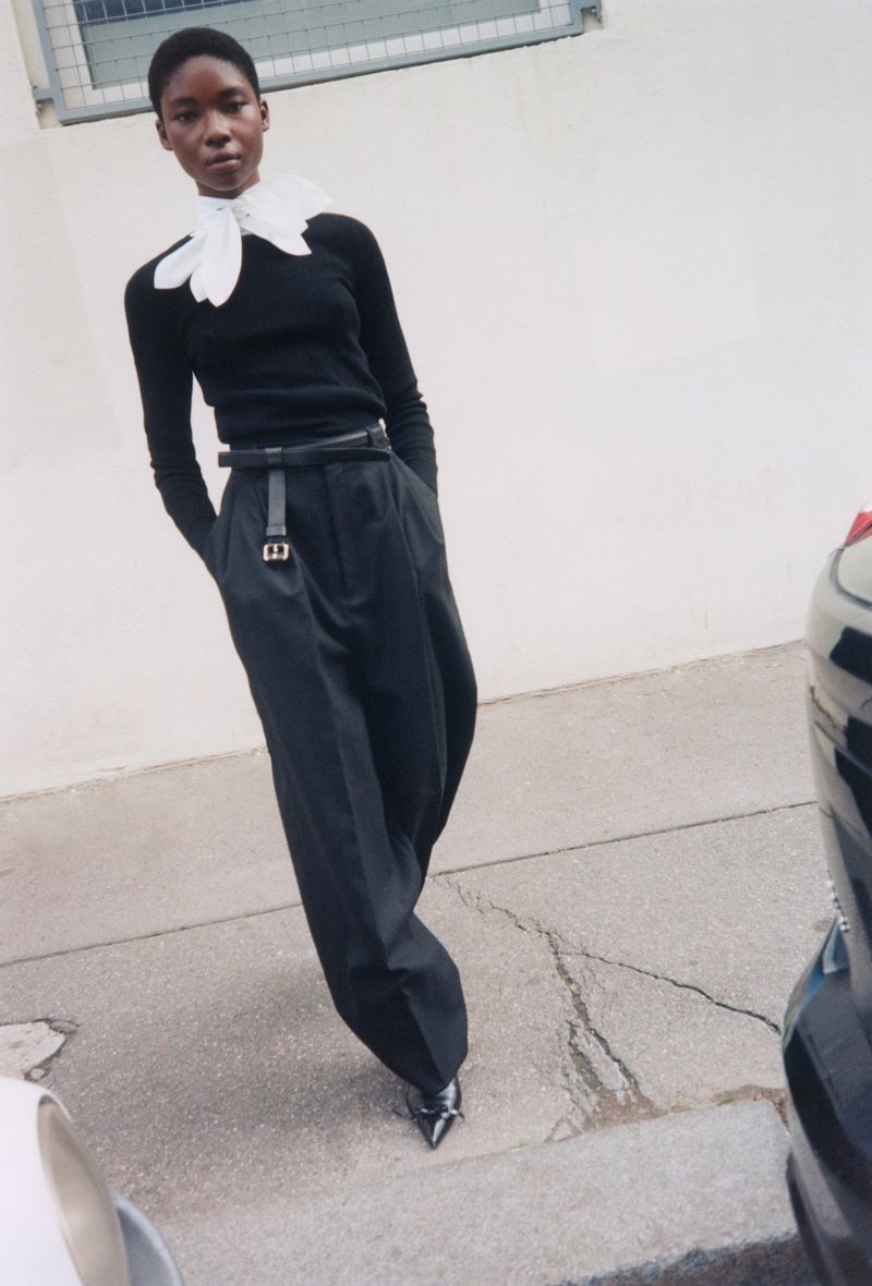 Elegant Black model in a studio-lit fashion shot, wearing a white collar, a black top, loose black trousers, and shiny shoes.