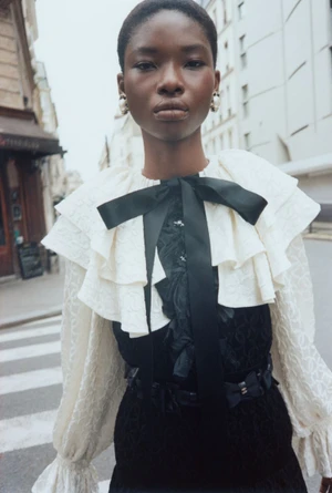 Black female model wearing a cream ruffled top with a black ribbon bow outdoors on a city street.