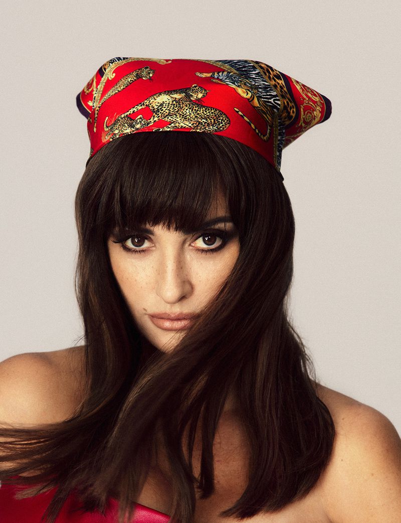 Portrait of an adult woman wearing a red patterned headscarf and makeup, posing in a studio setting with diffused lighting.