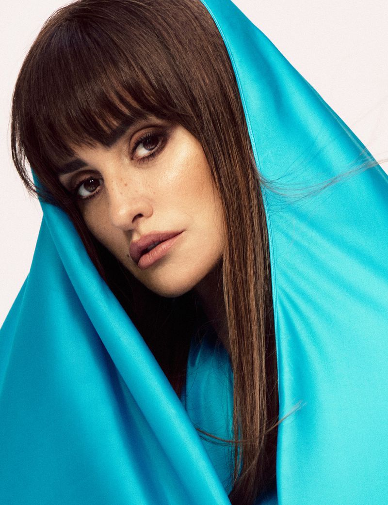 Close-up portrait of a brunette adult woman with bangs, framed by a draped piece of bright turquoise fabric against a neutral background.
