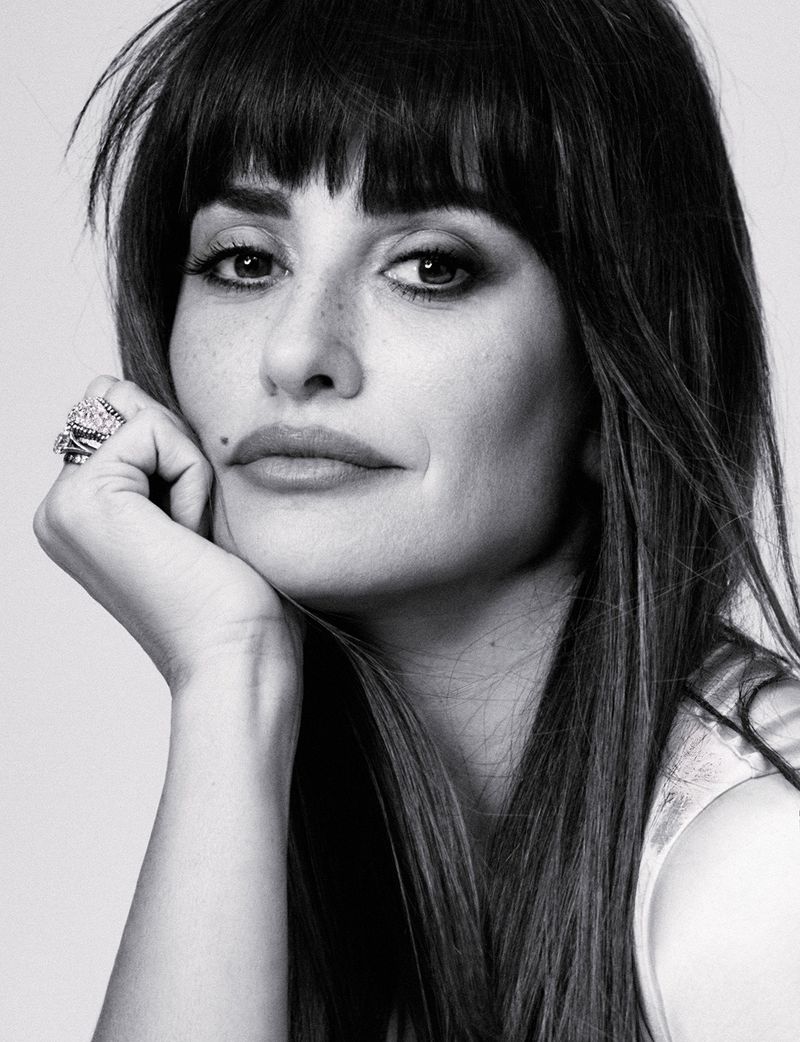 Black and white portrait of a woman with dark hair and bangs, resting her chin on her hand. She is wearing rings and has a mole on her face.
