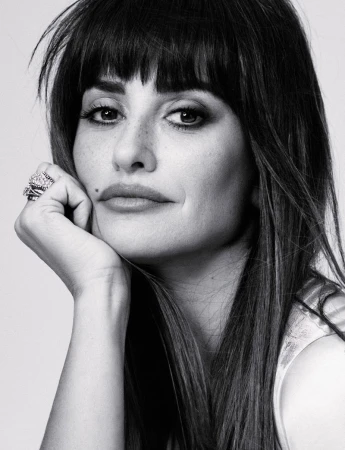Black and white portrait of a woman with dark hair and bangs, resting her chin on her hand. She is wearing rings and has a mole on her face.
