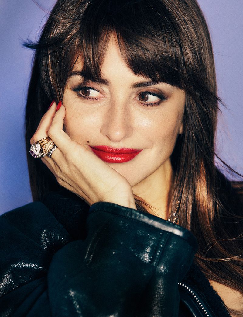 Close-up portrait of an adult female with dark hair, red lipstick, and rings, resting her chin on her hand against a purple backdrop.