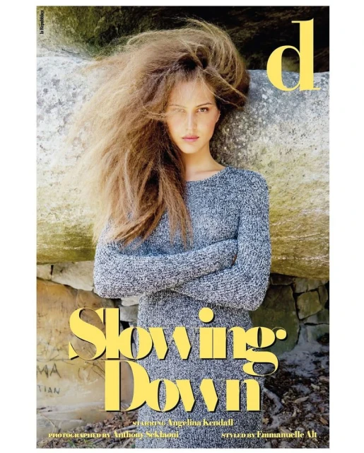 A young adult woman with long brown hair poses in a grey knit sweater dress in front of a stone wall in a medium portrait shot.