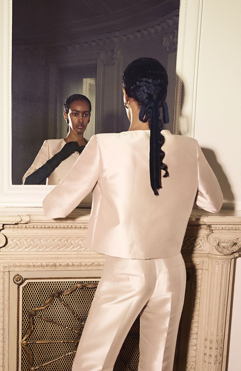 A woman in a pink pantsuit stands in front of a fireplace, her reflection visible in the mirror behind her. Fashion editorial.