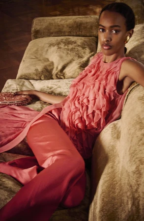 Model Amina Ahmed in a coral pink ruffled silk sleeveless top and matching trousers reclining on a plush velvet sofa.