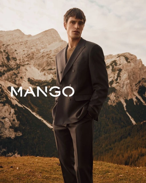 A confident adult male model in a dark suit stands outdoors against a backdrop of mountains for a Mango fashion advertisement.