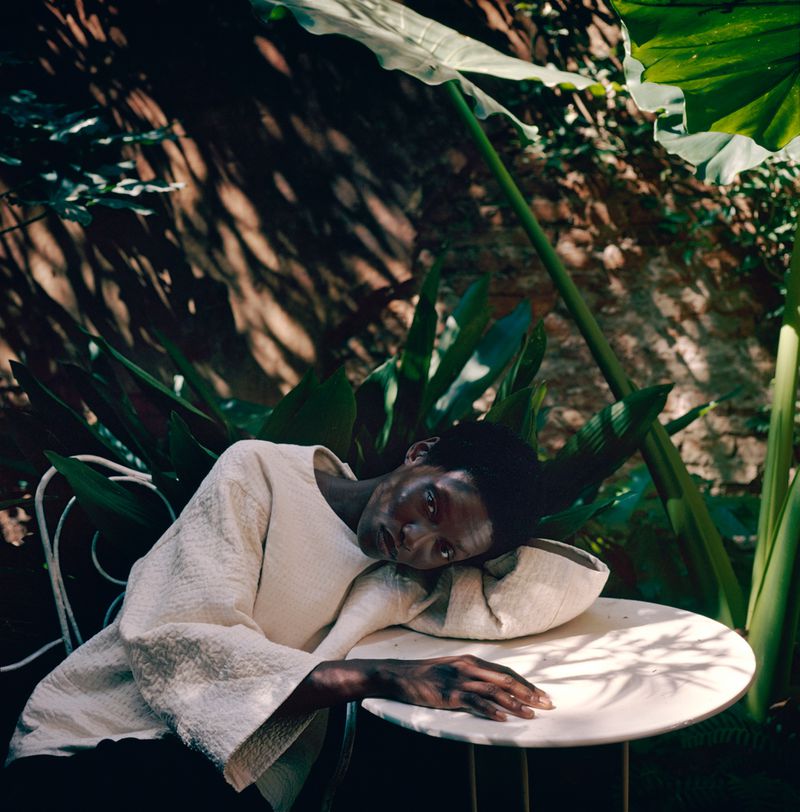 Woman rests head on table outdoors surrounded by greenery. Cream top, round table, dappled sunlight, dark hair, garden wall.