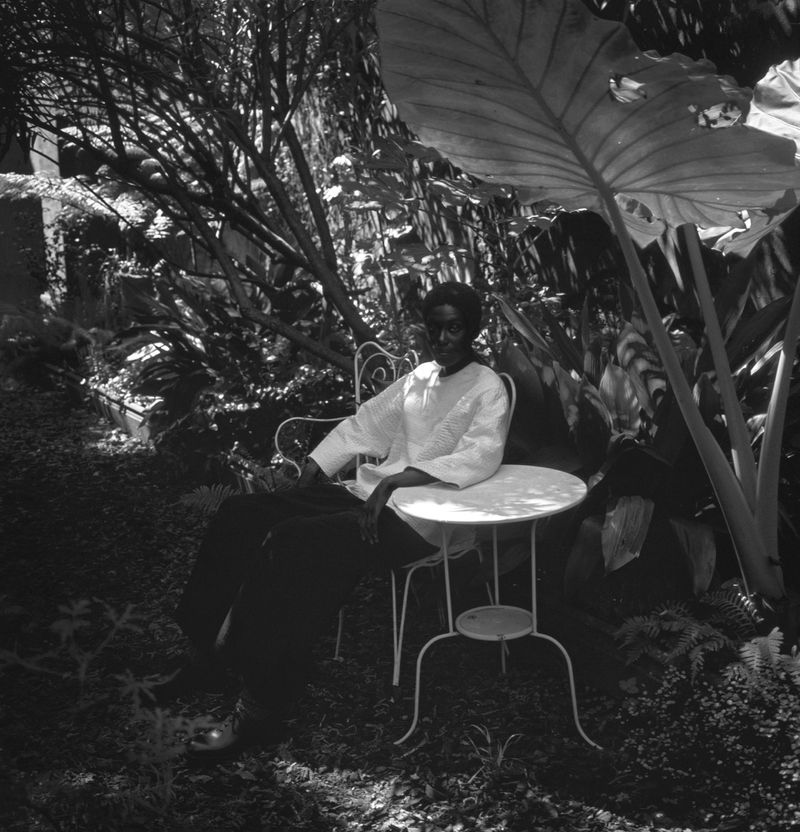 A black and white outdoor shot of a woman in a garden sitting at a small, round table under the shade of a large plant leaf.