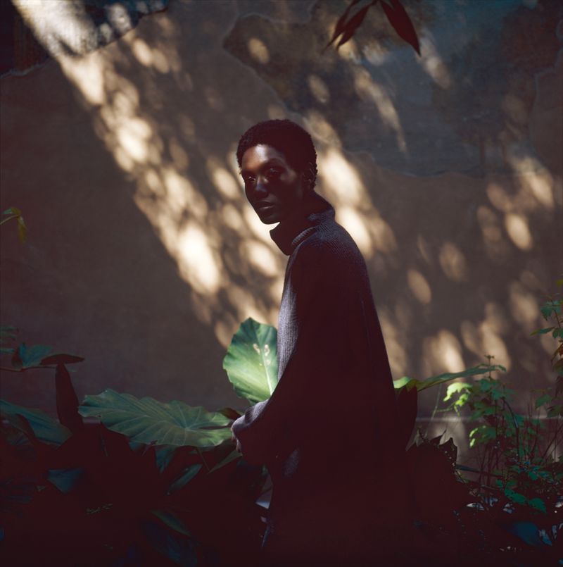 Dark-skinned adult stands in a natural setting with a blurred background and dappled light, wearing a dark collared coat.
