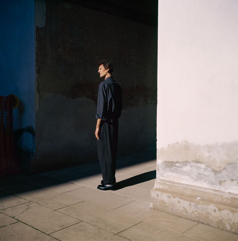Full-length shot of a slender adult man in a dark shirt and pants standing sideways on a stone floor between stark walls.