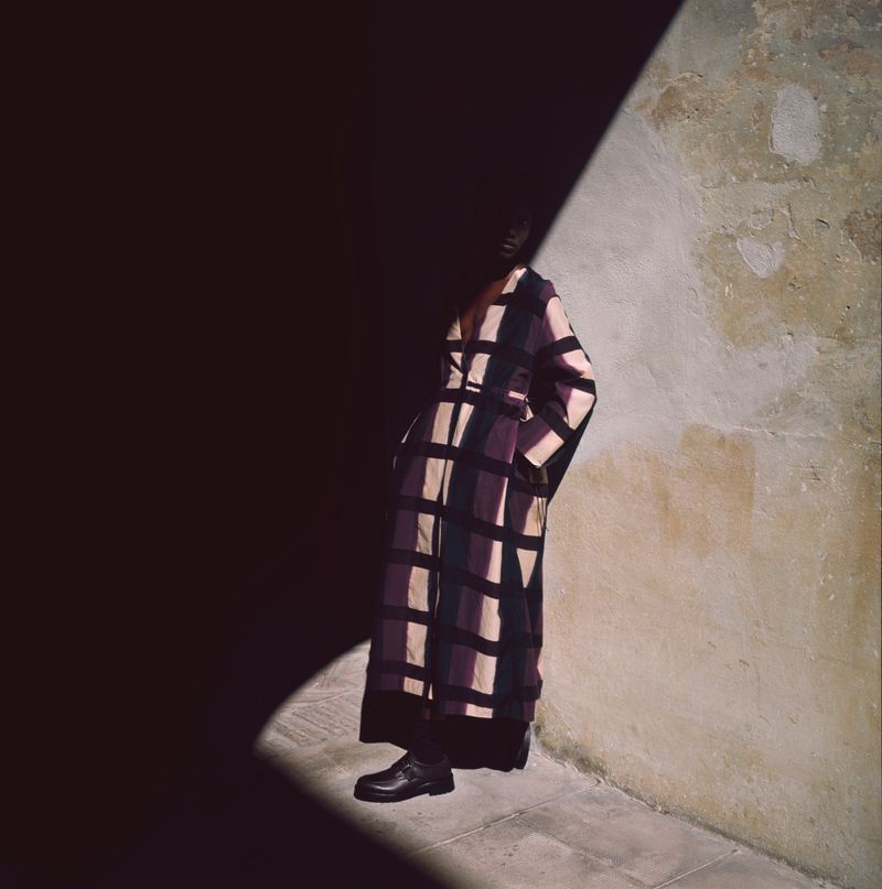 A person stands in a shadowy corner wearing a patterned brown and purple dress with black shoes next to a textured wall.