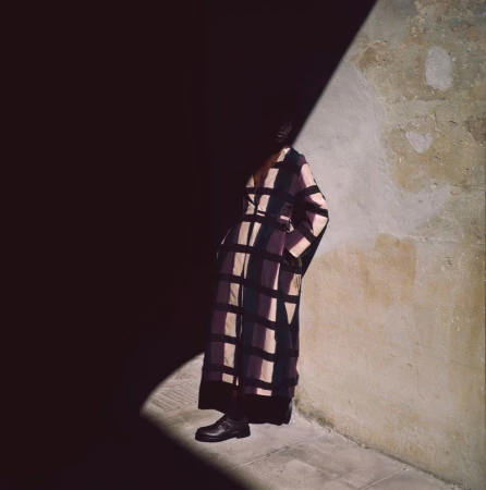 A person stands in a shadowy corner wearing a patterned brown and purple dress with black shoes next to a textured wall.