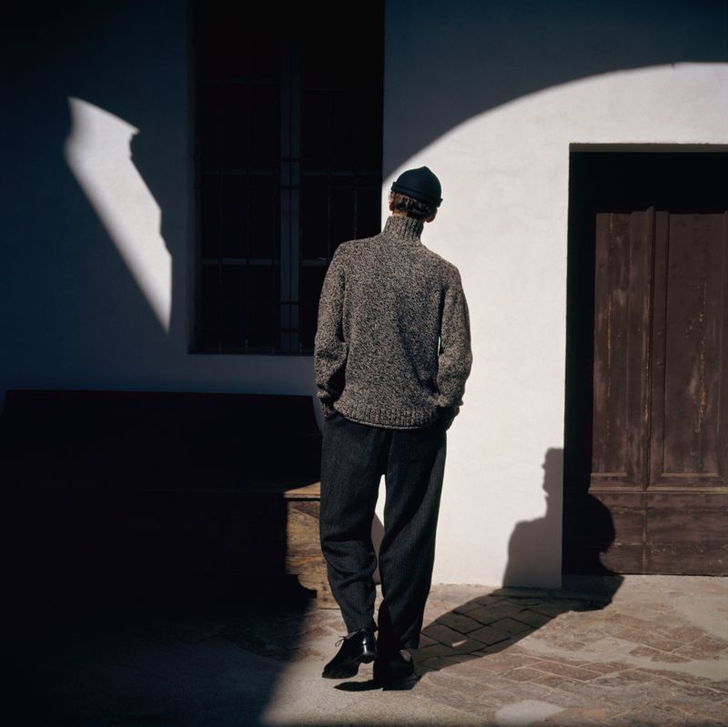 A man stands in a courtyard next to a building in a knit sweater, dark pants and a beanie hat, looking toward an arched doorway.