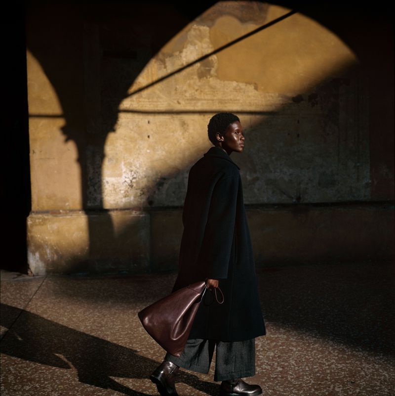A black model walks through an arched space with shadows, carrying a dark brown leather handbag and wearing a black coat and dark grey pants.