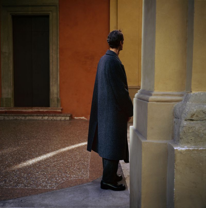 A man in a dark overcoat stands on a sunlit stone surface in an urban setting. The architecture features orange and yellow walls.