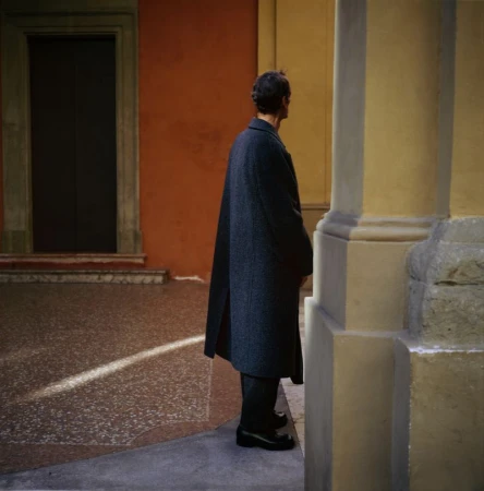 A man in a dark overcoat stands on a sunlit stone surface in an urban setting. The architecture features orange and yellow walls.