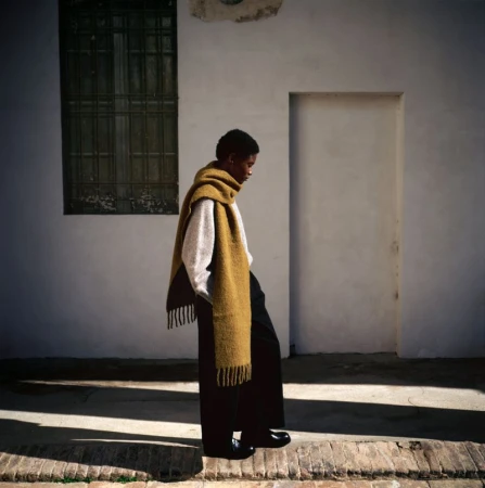 An adult model with short hair walks along a brick sidewalk wearing black pants, a white sweater and a large yellow scarf.