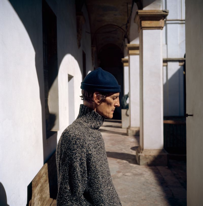 Adult male in gray turtleneck sweater and blue knit cap stands outside on a colonnade with columns and sunlight casting shadows.